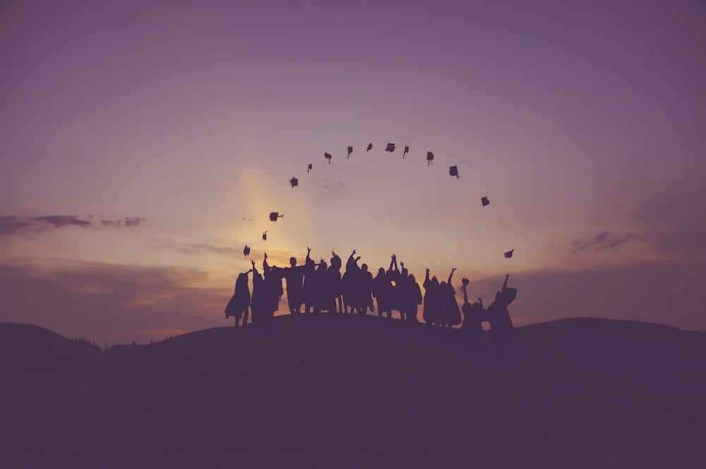 A row of graduates on a hill throwing their hats in a perfect rainbow shape.