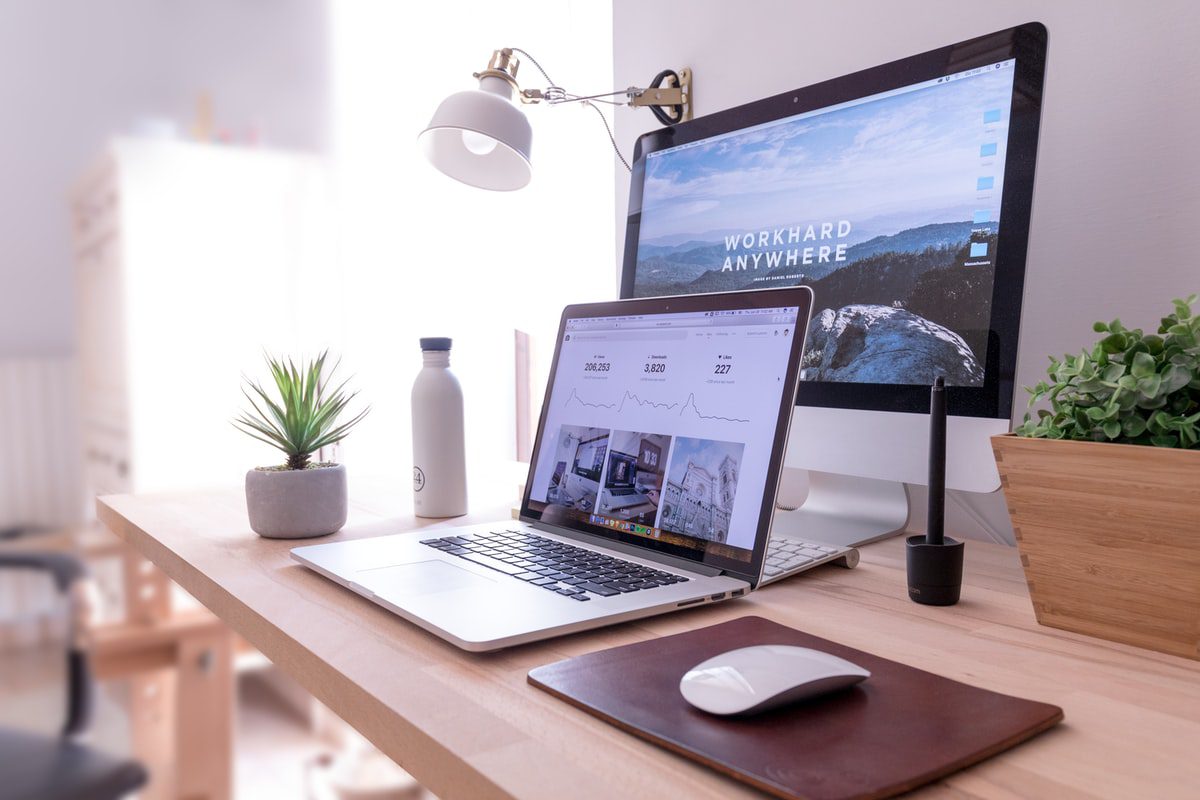A MacBook Pro, iMac, and Magic Mouse on a table next to two plants, a lamp, and a water bottle.